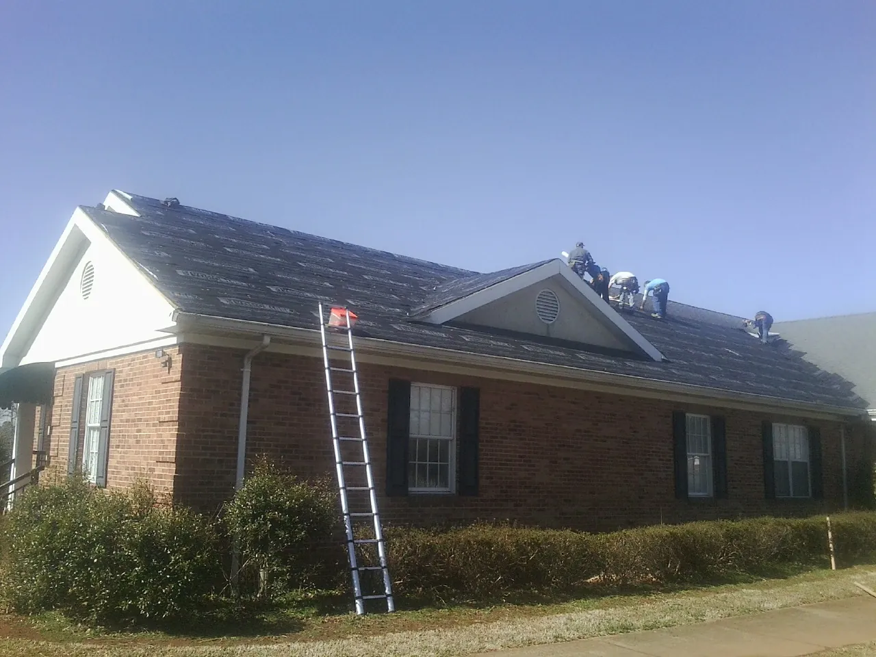 Crew tearing shingles off a brick ranch roof with a ladder set — Metal Roof Repair work by our crew in Lees Summit