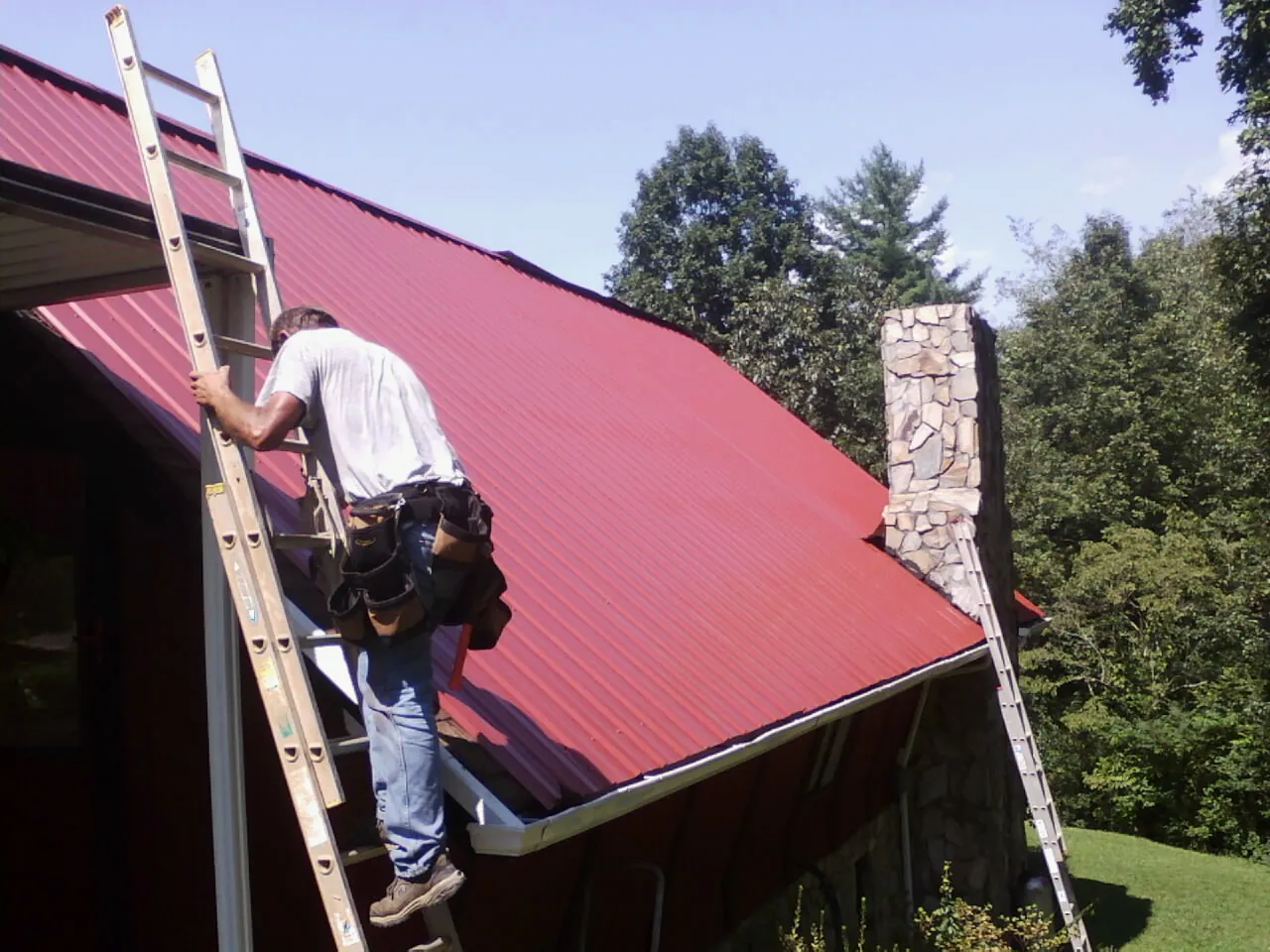 Roofer on a ladder beside a red metal roof and stone chimney — Soffit & Fascia Repair by Roofing Brothers in Lees Summit